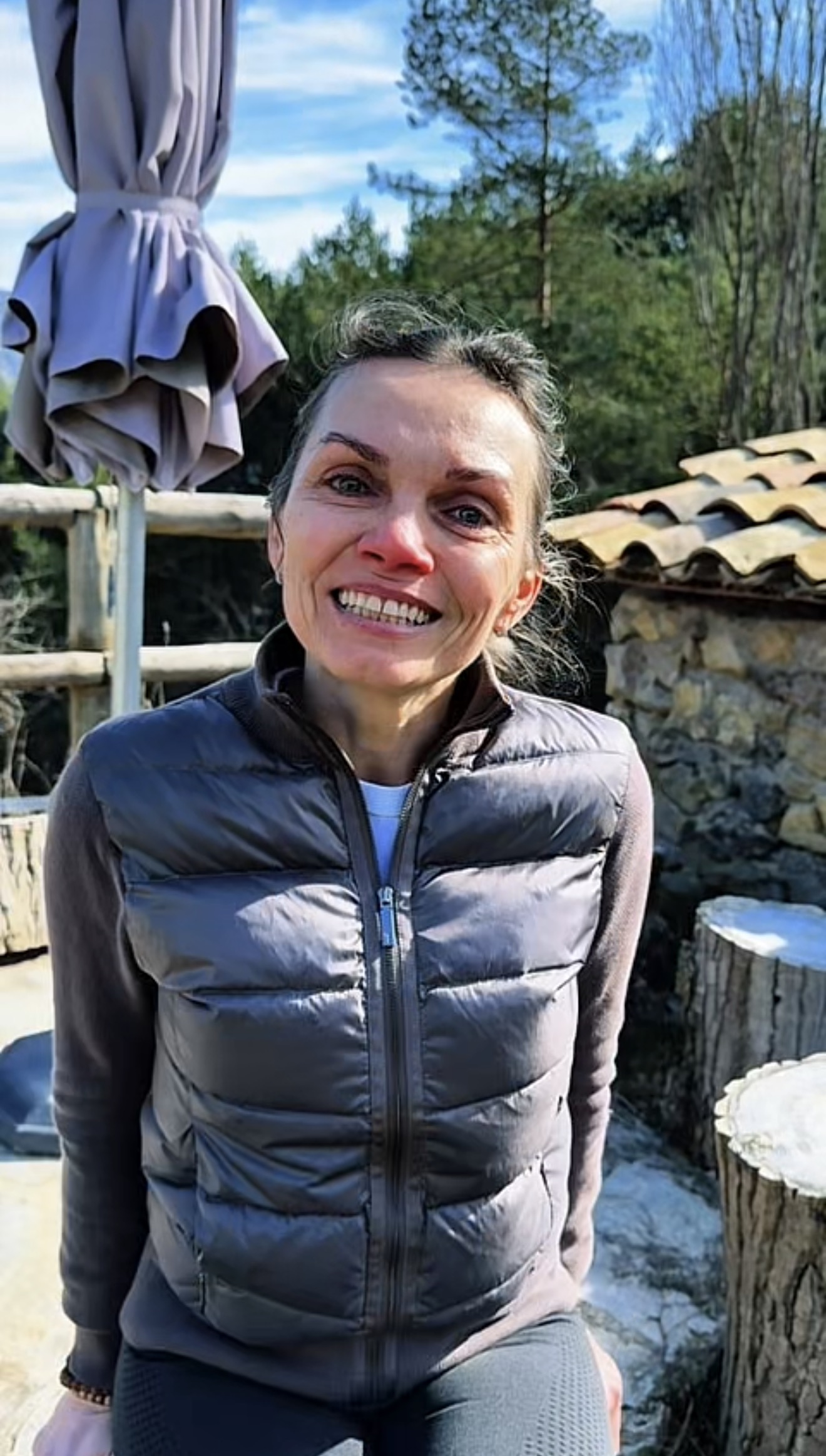 Spokojená klientka RE·CORDIS — usměvavá žena v šedé prošívané bundě sedí venku u kamenné zdi s stromy v pozadí
