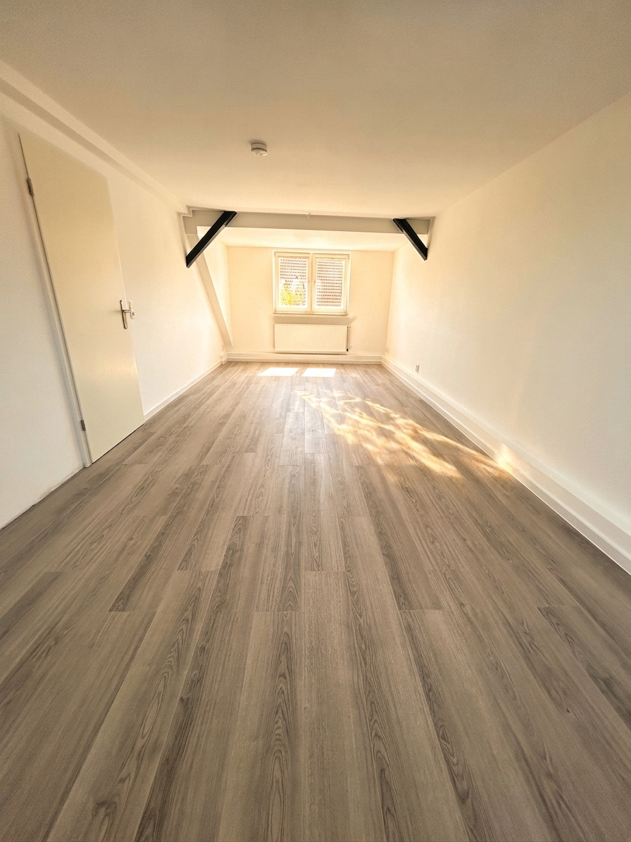 Helles Dachgeschosszimmer mit dunklen Holzbalken, Fenster und grauem Laminatboden – Dachgeschosswohnung Dessau