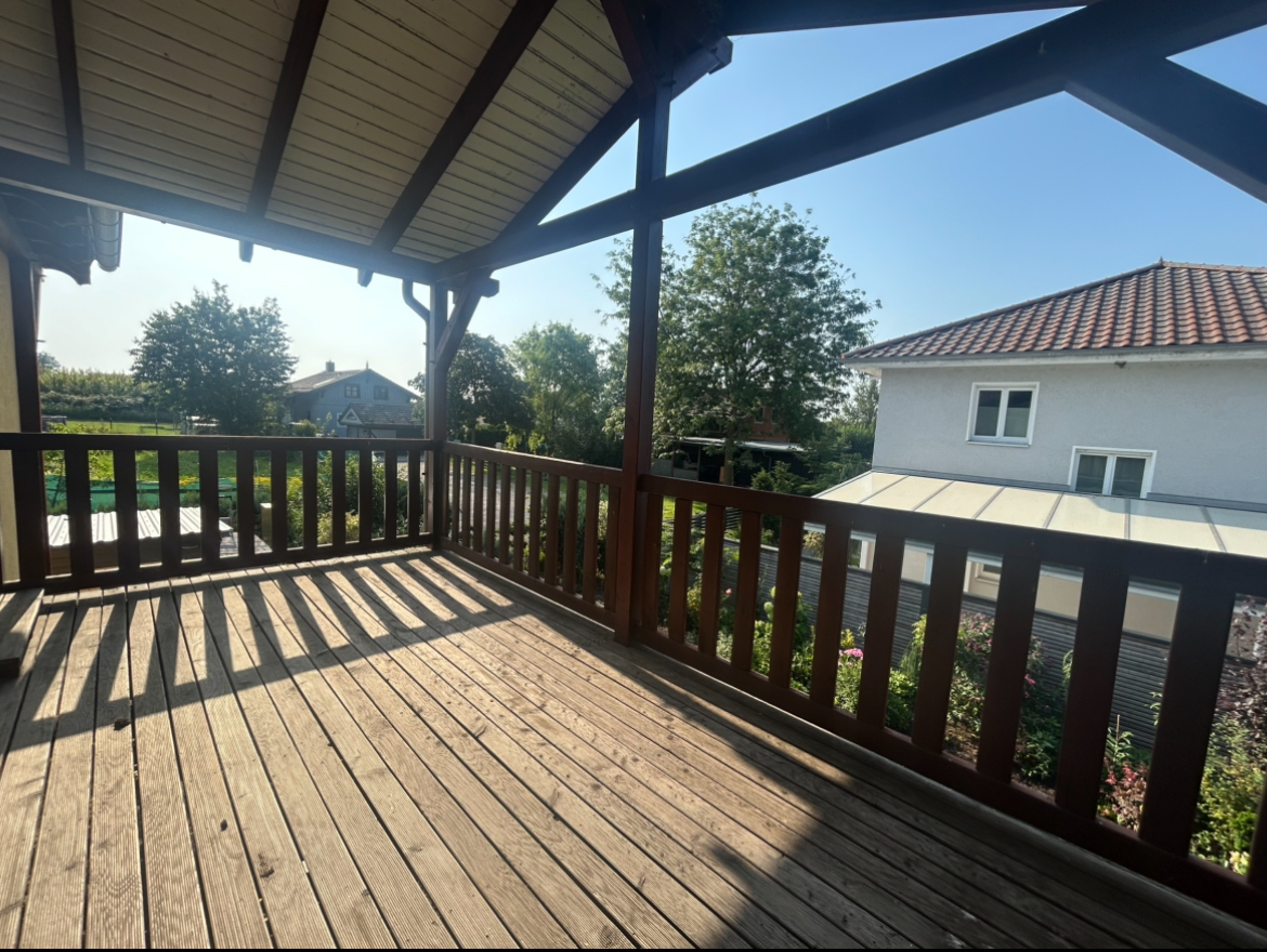Großer überdachter Balkon mit Holzdielen, Holzgeländer und Blick in den Garten – 3-Zimmer-Wohnung in Ketzin