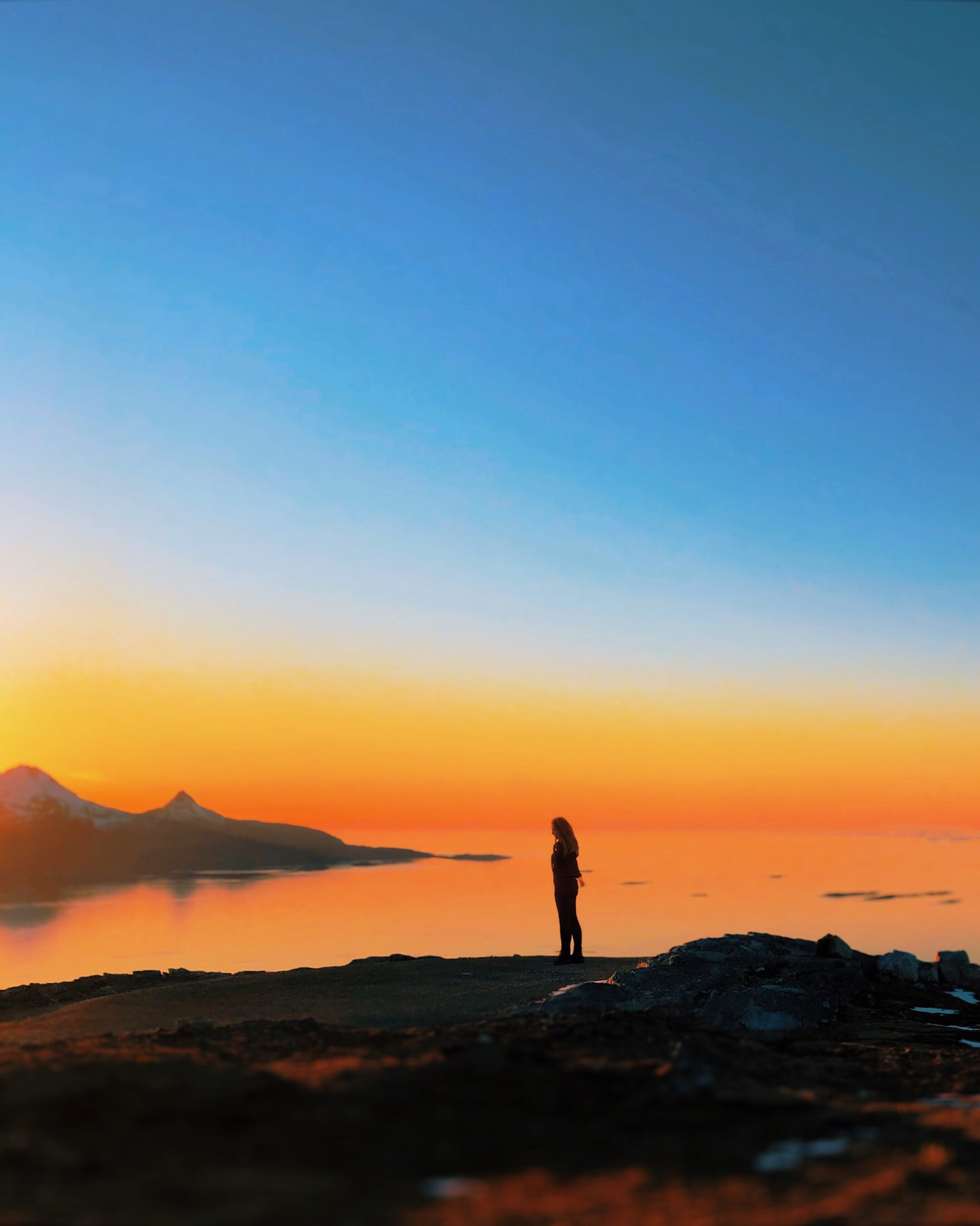 Silhuett av en kvinne som står alene på en fjelltopp og ser utover et stille hav i solnedgang, med en dramatisk oransje og blå himmel