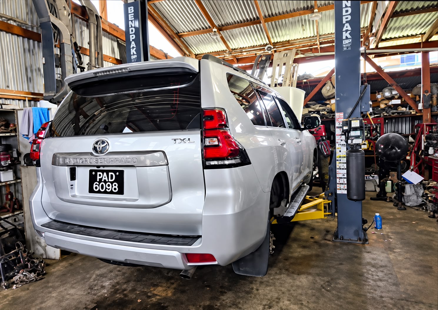 White Toyota Land Cruiser Prado TXL on a BendPak hydraulic lift inside L.Singh Mechanic Shop, rear view showing the full workshop bay