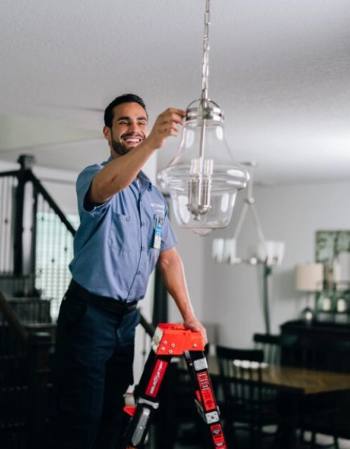 Smiling Mr. Electric technician on a ladder installing a light fixture in a home